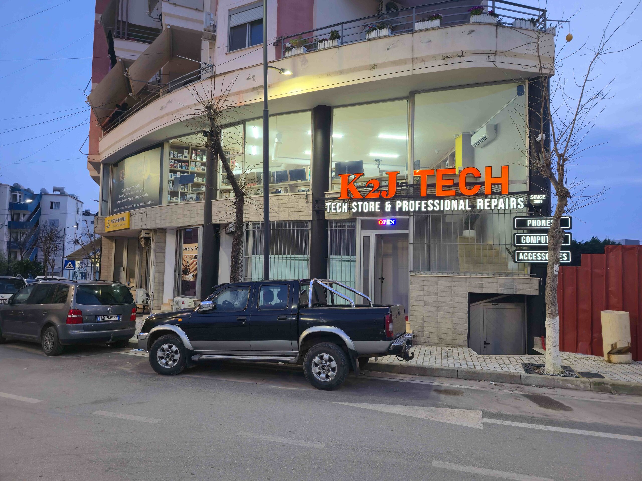 K2J Tech storefront illuminated at night in Sarandë, Albania showing the iconic orange signage